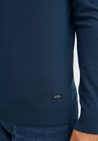Tamsiai mėlynas megztinis su raukšlėtais rankogaliais ir apačia. Ant apačios yra maža juoda etiketė su užrašu "SKIRTA DENIM PERFEKCIONUI."