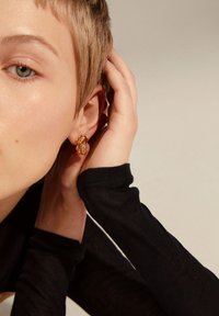 Orecchini con texture dorata e design intricato, indossati a un orecchio, abbinati a una maglietta nera a maniche lunghe. La tonalità della pelle appare neutra.