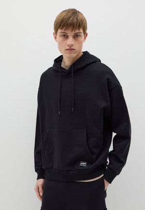 Sudadera con capucha negra fabricada en una mezcla de algodón. Presenta un bolsillo frontal, capucha con cordón y puños acanalados con una pequeña etiqueta de logo.