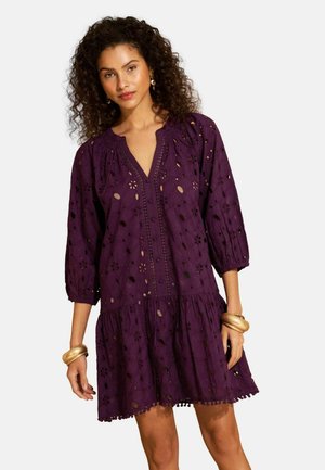 Kvinde iført en lilla øjetskjole med 3/4-ærmer, V-hals og en flæset kant, accessoriseret med guldbøjler og øreringe.