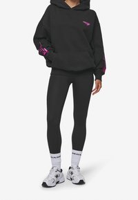 Černá mikina s kapucí a růžovými akcenty, kombinovaná s černými legínami a bílými teniskami. Obsahuje prostornou kapsu a volný střih.