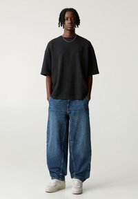 T-shirt noire en coton surdimensionnée, pantalon en denim bleu décontracté avec des détails de couture subtils et baskets blanches. Design simple et coupe décontractée.