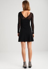 Czarna sukienka z długimi przezroczystymi rękawami z wzorami gwiazd, dopasowaną górą i rozkloszowanym dołem. Tył ma dekolt w kształcie litery V.