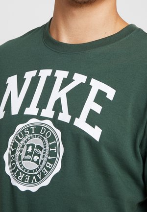 Mężczyzna w ciemnozielonej koszulce Nike z białym logo i okrągłym emblematem z napisem „Winning Spirit, Since 1972, Beaverton, Just Do It.”