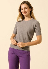 Pull gris à manches courtes avec un col rond, en matériau tricoté et à la coupe ajustée. Assorti avec un pantalon violet, sur un fond neutre.
