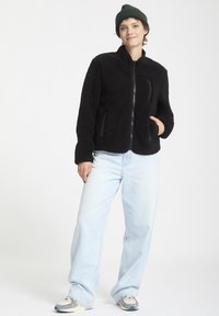 Giacca in fleece nera con colletto alto, chiusura a zip e tasche laterali, abbinata a jeans a gamba larga blu chiaro e un berretto verde.