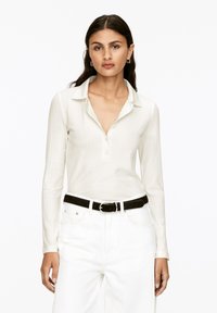 Young woman with long dark hair wears a ribbed cream shirt with collar, white pants, and a black belt, standing against a white background.