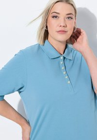 Světle modrá polo tričko s krátkými rukávy, límečkem a zapínáním na pět knoflíků. Měkká, strukturovaná tkanina s volným střihem.