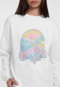 Sudadera blanca con un gráfico colorido de olas, sol y palmeras, con el texto "Guess Jeans" en tonos rosa y azul.