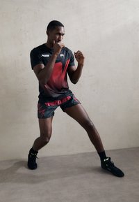 Schwarzes Sportshirt mit rotem Grafikdesign, kombiniert mit schwarzen Shorts mit roten Blitzakzenten. Schwarze Sportschuhe mit gelben Details.