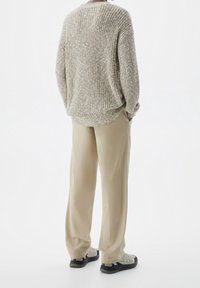 Personne debout, les mains dans les poches, portant un pull en maille beige, un pantalon kaki ample et des baskets grises sur un fond blanc.