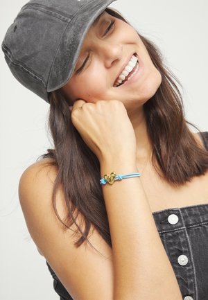Joven sonriente con los ojos cerrados, lleva una gorra gris, una blusa negra abotonada y una pulsera azul con un dije de corazón dorado.