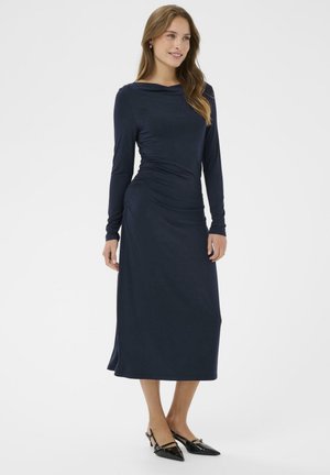 Navy Langarmkleid mit drapiertem Ausschnitt und gerüschtem Seitendetail, aus weichem Stoff gefertigt, kombiniert mit schwarzen spitzen Pumps.