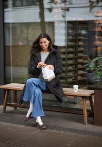 Schwarze Daunenjacke mit Kapuze, gestreiftes Hemd, blaue weite Jeans, braune Sneaker, hält ein Gebäck in einer Papiertüte, Kaffeebecher in der Nähe.