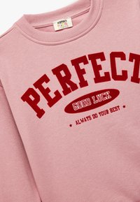Vaaleanpunainen huppari, jossa on punainen teksti "PERFECT", "GOOD LUCK" ja "ALWAYS DO YOUR BEST". Pehmeää kangasta, ribbauskaulus ja rento muotoilu.