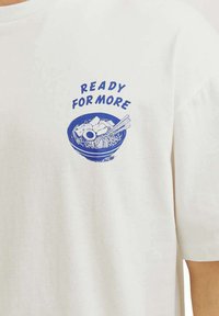 T-shirt bianco in cotone con grafica blu di una ciotola di noodle con il testo "PRONTO PER ALTRO" sopra. Maniche corte, vestibilità rilassata.
