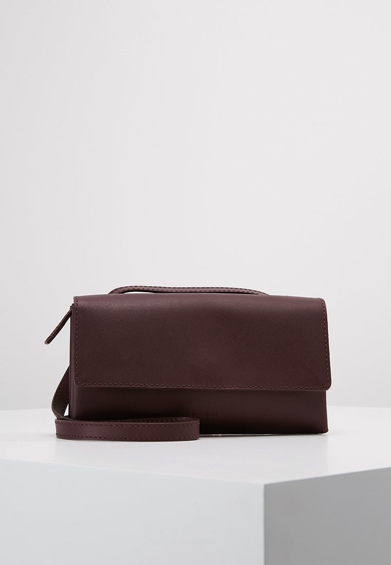Hnědá kožená crossbody taška s klopovým uzávěrem a nastavitelným popruhem, položena na bílem podstavci na jednobarevném pozadí.