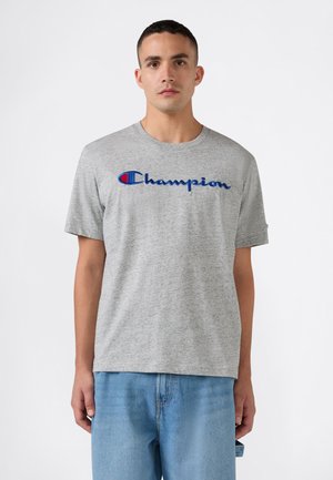 Man die een grijs Champion T-shirt met blauw logo en lichtblauwe spijkerbroek draagt, staand tegen een witte achtergrond.