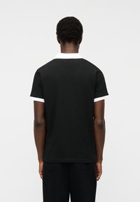 Άτομο με κοντά σγουρά μαλλιά, που φοράει μαύρο κοντομάνικο πουκάμισο με λευκό γιακά και άκρες στα μανίκια, φαίνεται από πίσω με φόντο ένα απλό υπόβαθρο.