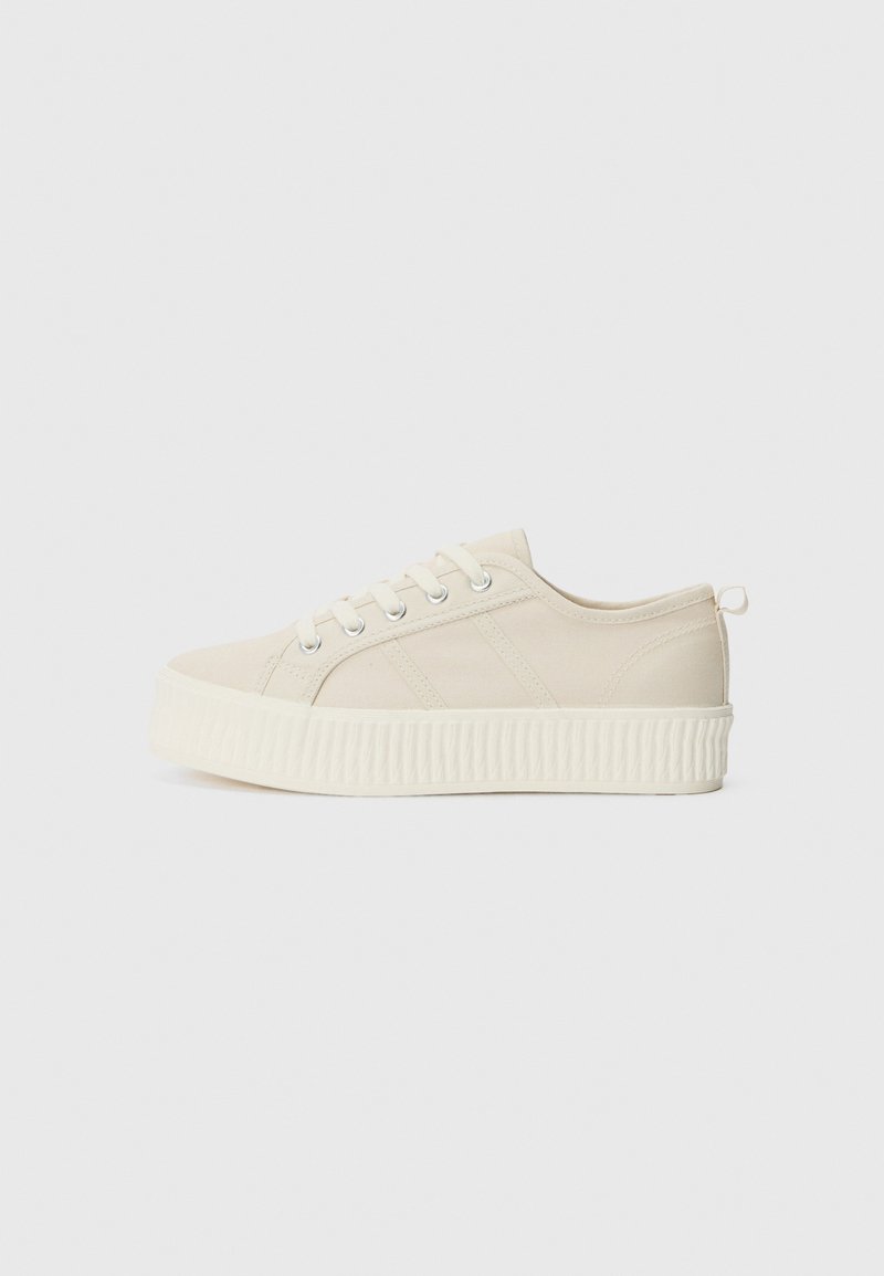 Sneaker in tela beige con suola bianca spessa e rigata, punta rotonda e design a tre strisce. Presenta occhielli in metallo e un tab di supporto sul tallone.