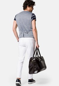 Brązowa skórzana torba podróżna z podwójnymi uchwytami, zamknięciem na zamek i gładką teksturą, zestawiona z szarym w kratkę kamizelką i białymi spodniami.