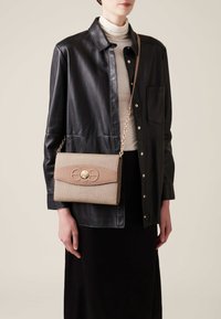 Beige textured handbag with a smooth tan flap, gold hardware, and chain strap. Model wears a black leather shirt jacket and a cream turtleneck.