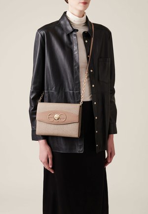 Sac à main beige texturé avec un rabat en cuir lisse couleur tan, des accessoires en or et une chaîne. Le modèle porte une veste en cuir noire et un col roulé crème.