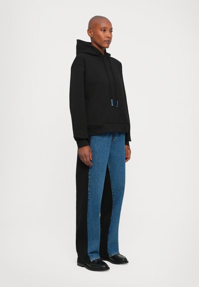 Schwarzer Kapuzenpullover mit lockerer Passform. Kombiniert mit Denim-Hosen mit geteilten Beinen, die eine blaue Vorderseite und schwarze Seitenpaneele aufweisen. Schwarze Schuhe.