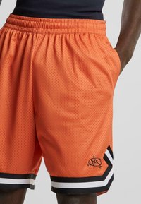 Orange netshorts med elastisk talje, sidelommer og sort-hvide striber ved kanten. Har et logo-detalje.