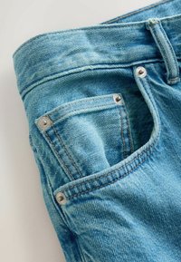 Gros plan sur la poche d'un jean en denim bleu clair avec des coutures visibles, des rivets métalliques et la texture du tissu sur un fond clair.