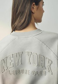 Világosszürke pulóver, amelyen a "NEW YORK LEISURE CLUB" felirat hímzett, ellentétes krémszínben. Ribbed nyak és raglánujjak.