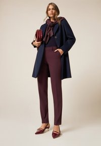 Marinejas, bordeaux kleurige sjaal, aansluitend zwart topje en op maat gemaakte bordeaux kleurige broek. Gepunte gepatineerde schoenen met een bandje. Gestructureerde handtas inbegrepen.