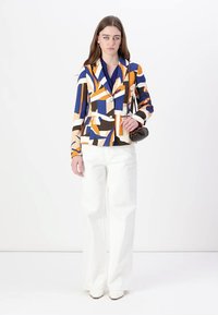 Blazer multicolore con motivi geometrici in blu, arancione e marrone, dotato di revers a punta, due bottoni e pantaloni bianchi a gamba larga.