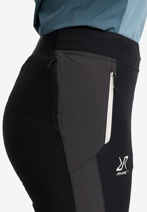 Närbild av svarta och grå idrottsbyxor med dragkedjefickor och RVRC-logotyp på låret, bärs tillsammans med en blå topp.