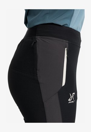 Närbild av svarta och grå idrottsbyxor med dragkedjefickor och RVRC-logotyp på låret, bärs tillsammans med en blå topp.