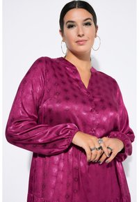 Magenta, satin Hemd mit langen Puffärmeln und einem geknöpften V-Ausschnitt. Es hat ein strukturiertes Muster und eine lockere Passform, die bis zur Hüfte reicht.