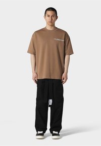 Brun oversized t-skjorte med hvit logo tekst, parret med svarte vide bukser med knytbelte og et merkesymbolet. Svarte joggesko fullfører looken.