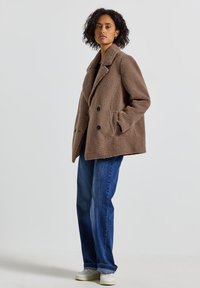 Kvinna med lockigt hår som bär en taupe dubbelknäppt teddyjacka, blå jeans och vita sneakers, står med händerna i fickorna.