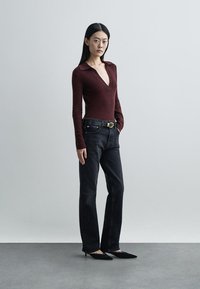 Γυναίκα όρθια, φορώντας μπορντό πουλόβερ με βαθύ V, μαύρα ψηλόμεσα τζιν με ζώνη και μαύρα μυτερά τακούνια.