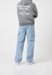 Harmaa huppari, jossa valkoinen teksti takana, yhdistetty vaaleansinisiin, väljisiin farkkuihin, joissa useita taskuja. Malli kasvot poispäin yksiväristä taustaa vasten.