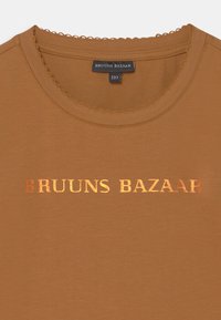 T-shirt de algodão castanho com decote redondo e costura decorativa em forma de onda. Apresenta o texto em dourado metálico "BRUUNS BAZAAR" na frente.