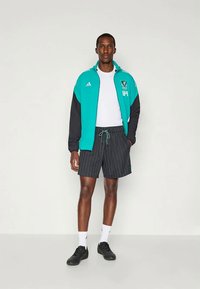 Chaqueta deportiva en azul verdoso y negro con logo, camiseta blanca, pantalones cortos negros a rayas y zapatillas negras con calcetines blancos.
