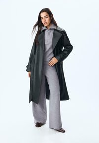 Schwarzer Leder-Trenchcoat über einem grauen Reißverschluss-Oberteil und passenden weiten Hosen. Glatte Textur, doppelt geknöpftes Design und Knöpfe. Braune Schuhe.