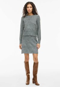 Suéter gris de punto y falda a juego, con un diseño texturizado. Botas marrones de tacón alto hasta la rodilla. Cuello redondo sencillo y silueta ajustada.