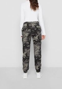 Pantalones cargo de camuflaje en tonos de gris y negro, con puños elásticos, bolsillos laterales y cremallera en la bragueta. Combinados con una camiseta blanca de manga larga.