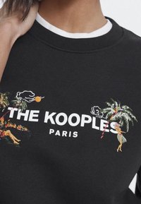 Černá mikina s bílým textem "THE KOOPLES PARIS." Na každé straně se nachází barevná výšivka palem a tropické scény.