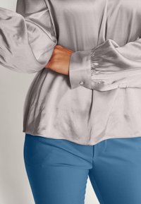 Blouse en satin gris clair avec manches bouffantes, ourlet arrondi et poignets à boutons, associée à un pantalon bleu ajusté et lisse.