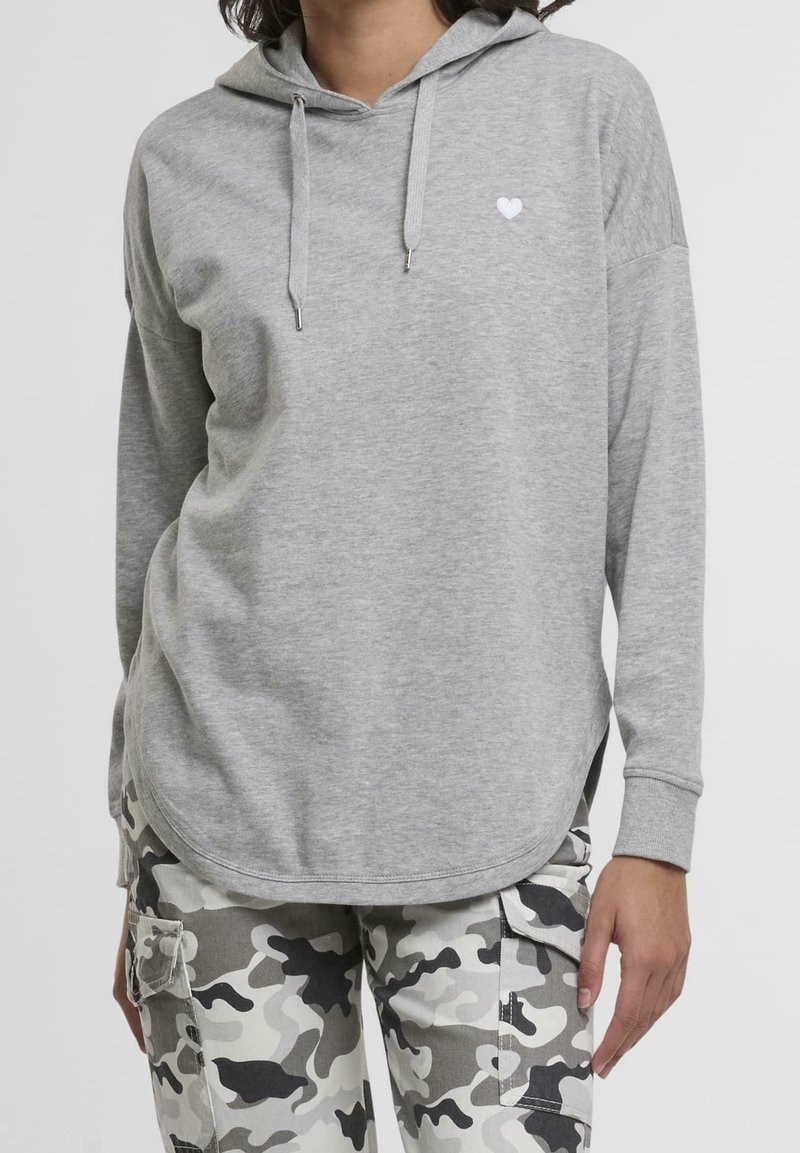 Personne portant un sweat à capuche gris clair avec un petit détail cœur blanc et un pantalon cargo camouflage noir, blanc et gris.