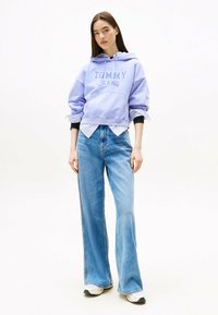 Felpa viola chiaro con stampa "TOMMY JEANS", indossata su una camicia a righe, abbinata a jeans larghi blu e sneakers bianche.
