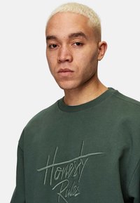 Grön sweatshirt med ribbad krage och broderad text "Ärlighet Regler" i en ljusare grön nyans. Tyget ser mjukt och strukturerat ut.